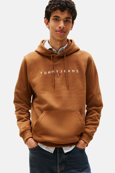 Bruine hoodie van TOMMY JEANS, met trekkoord en kangoeroezak, gedragen over een gestreept overhemd en een spijkerbroek.