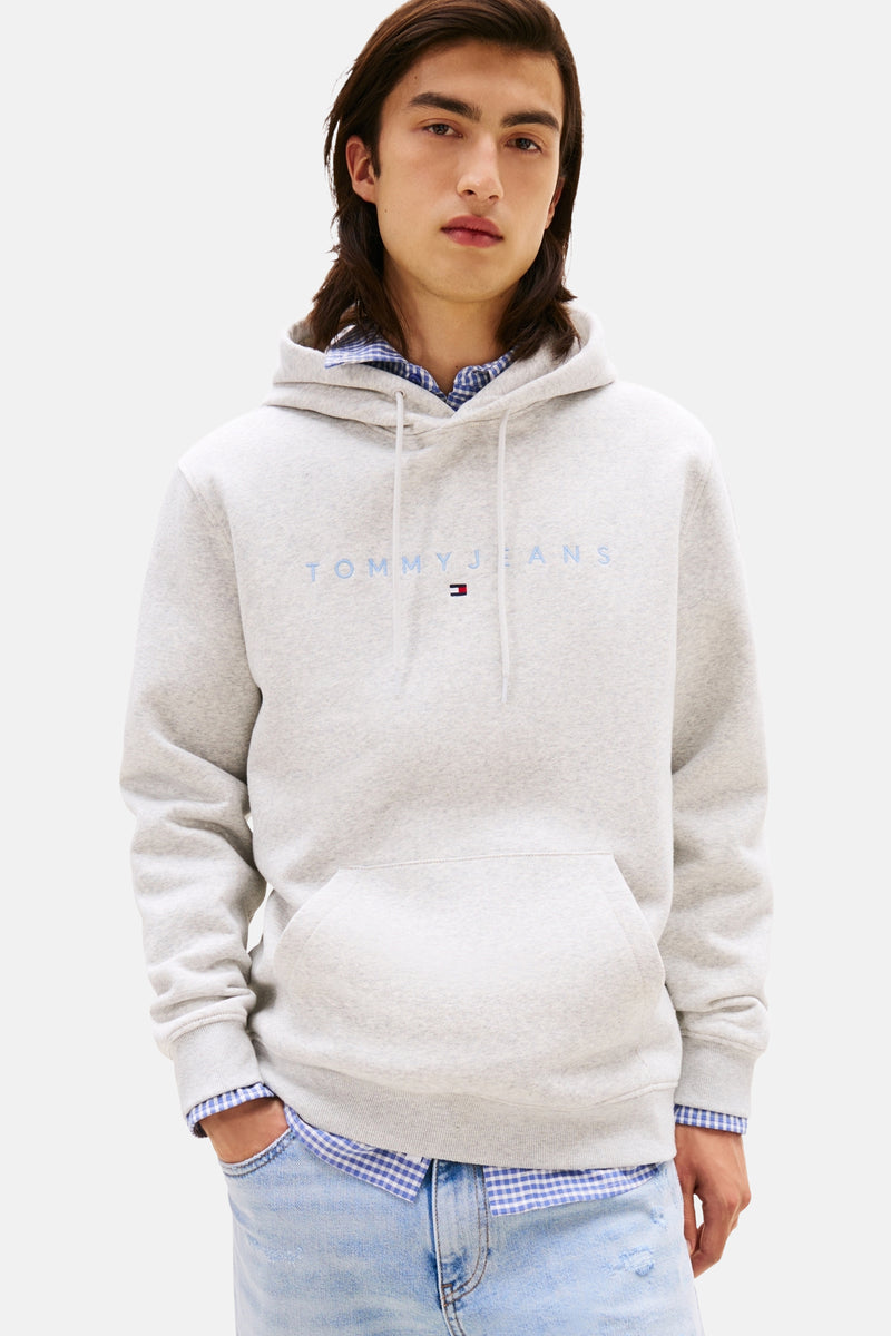 Grijze hoodie van TOMMY JEANS met trekkoord, gedragen over een geruit overhemd en een lichte spijkerbroek.