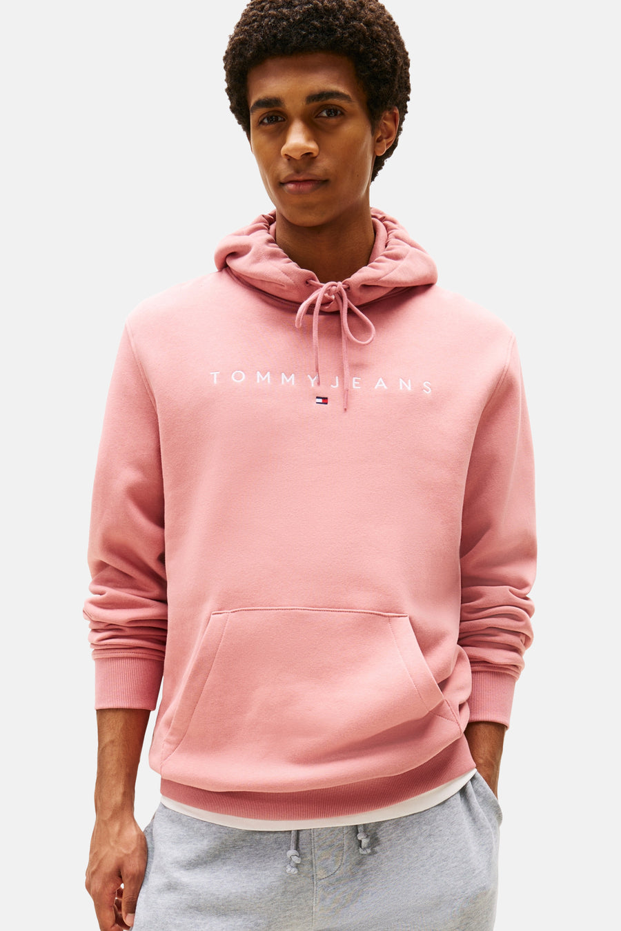 Sweat à capuche rose de TOMMY JEANS, avec cordon de serrage et poche kangourou, porté avec un pantalon de survêtement gris.
