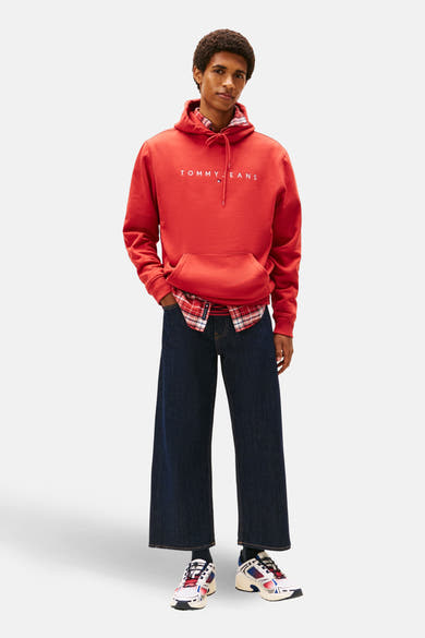 Sweat à capuche rouge par TOMMY JEANS porté avec une chemise à carreaux et un jean large.