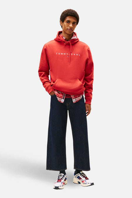 Sweat à capuche rouge par TOMMY JEANS porté avec une chemise à carreaux et un jean large.