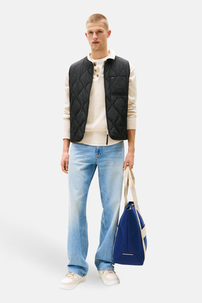 Veste sans manches noire de TOMMY JEANS, portée sur un pull boutonné crème avec un jean délavé, et tenant un sac bleu.
