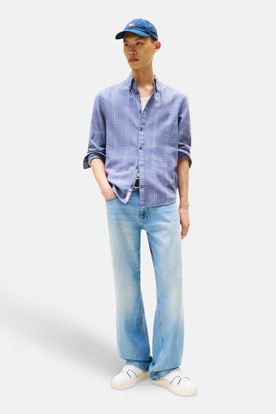 Chemise à manches longues bleue à carreaux de TOMMY JEANS, portée ouverte avec un t-shirt blanc, jeans bleu clair, casquette et baskets blanches.
