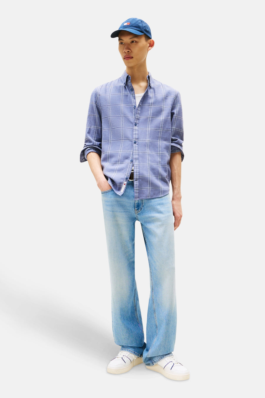 Chemise à manches longues bleue à carreaux de TOMMY JEANS, portée ouverte avec un t-shirt blanc, jeans bleu clair, casquette et baskets blanches.
