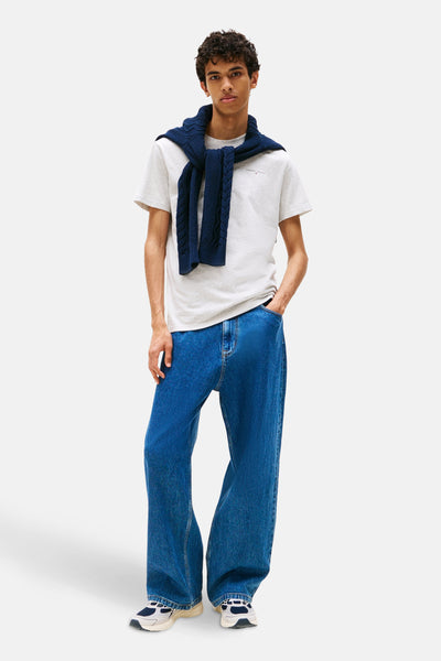 Grijs T-shirt met korte mouwen van TOMMY JEANS, gedragen met een marineblauwe trui over de schouders en een wijde blauwe jeans.