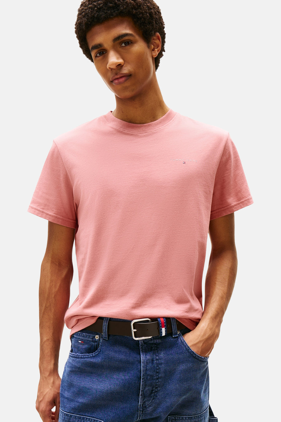 T-shirt rose à manches courtes de TOMMY JEANS, porté avec un jean bleu et une ceinture marron.
