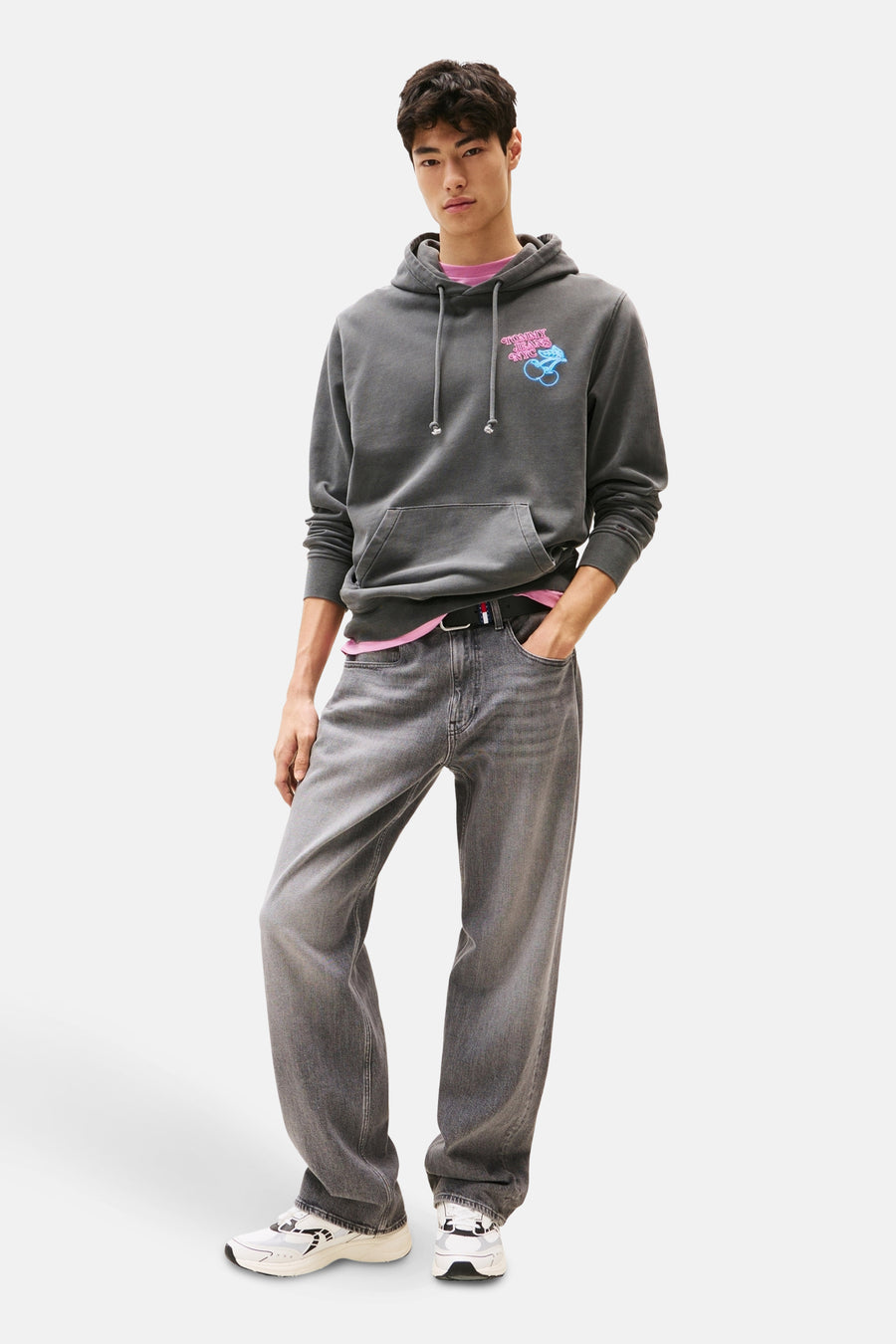 Sweat à capuche gris by TOMMY JEANS, avec un logo imprimé et une poche kangourou, porté avec un t-shirt rose et un jean.
