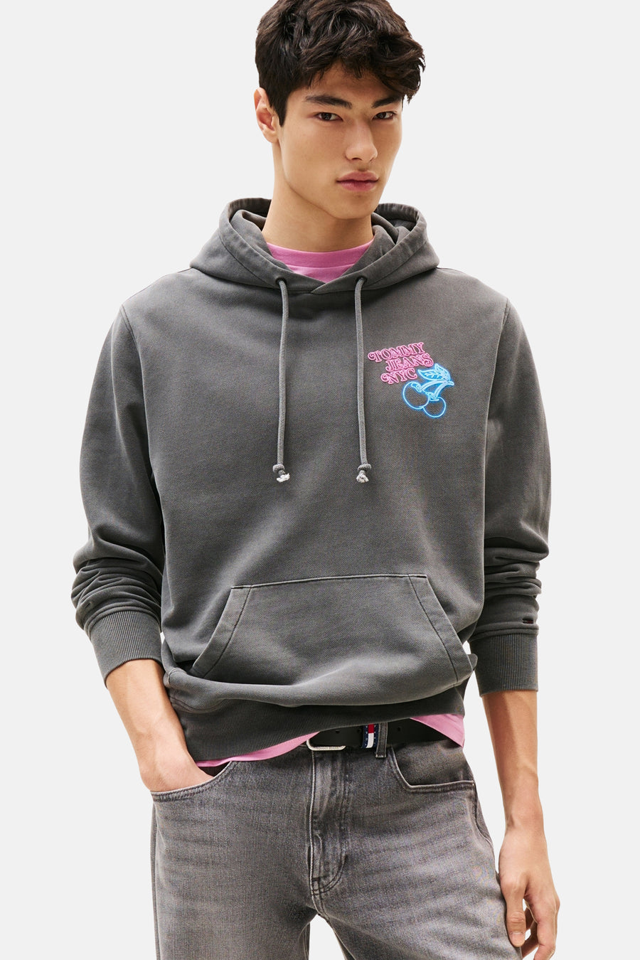 Sweat à capuche gris de Tommy Jeans, avec un logo imprimé, porté sur un t-shirt rose, avec un jean gris.
