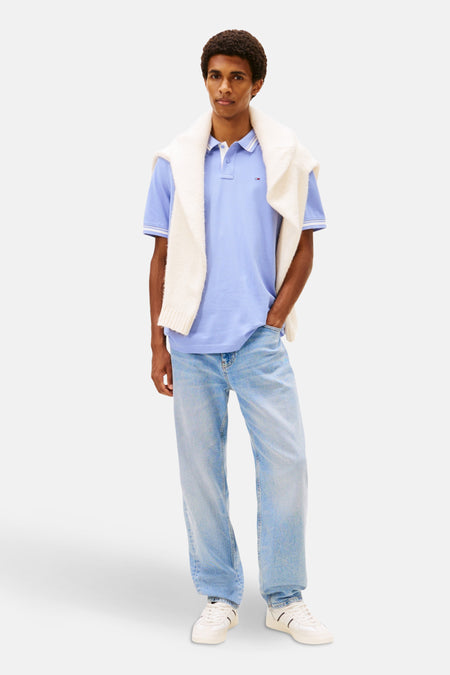 Polo mauve à manches courtes de TOMMY JEANS, porté avec un pull crème noué sur les épaules.