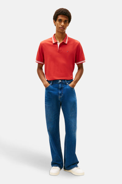 Rood poloshirt met korte mouwen van TOMMY JEANS, gedragen met een wijde blauwe jeans en witte sneakers.