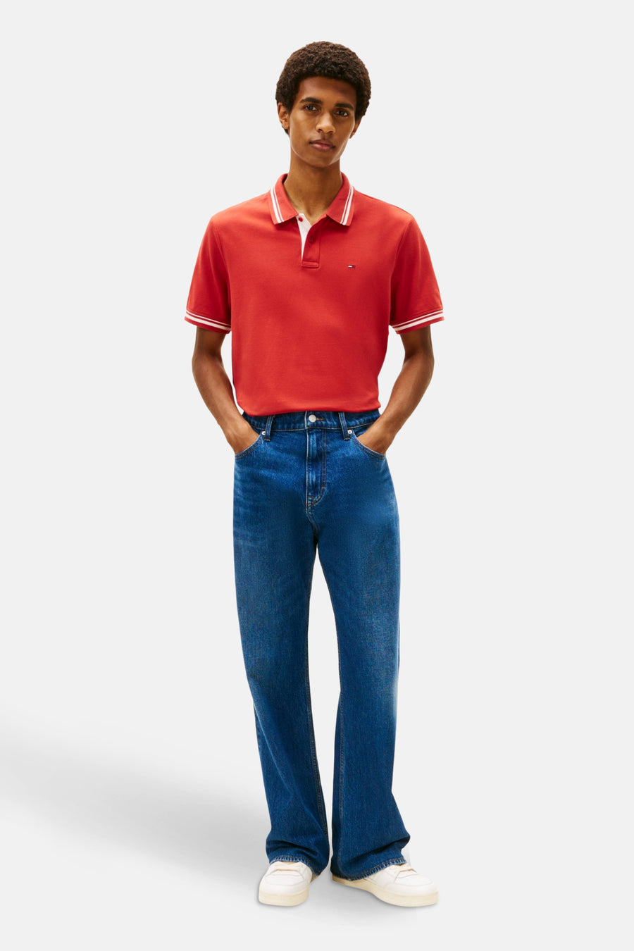 Rood poloshirt met korte mouwen van TOMMY JEANS, gedragen met een wijde blauwe jeans en witte sneakers.