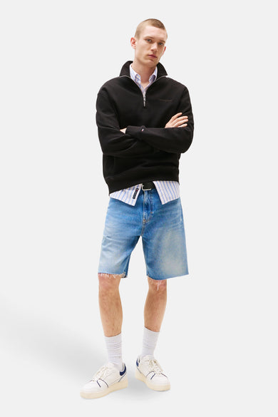Sweat noir de TOMMY JEANS, porté avec une chemise à rayures, un short en jean et des baskets blanches.