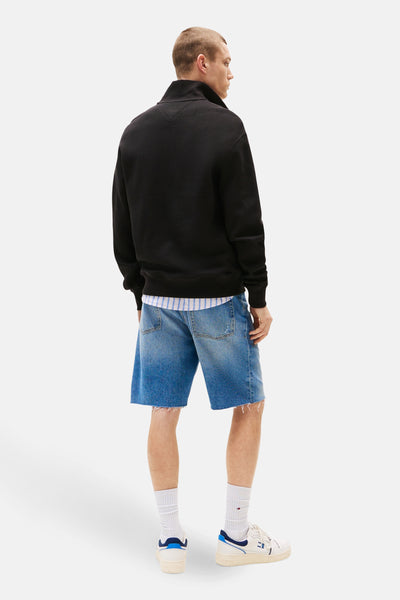 Sweat noir par TOMMY JEANS, texture gaufrée, porté sur une chemise rayée et un short en jean.