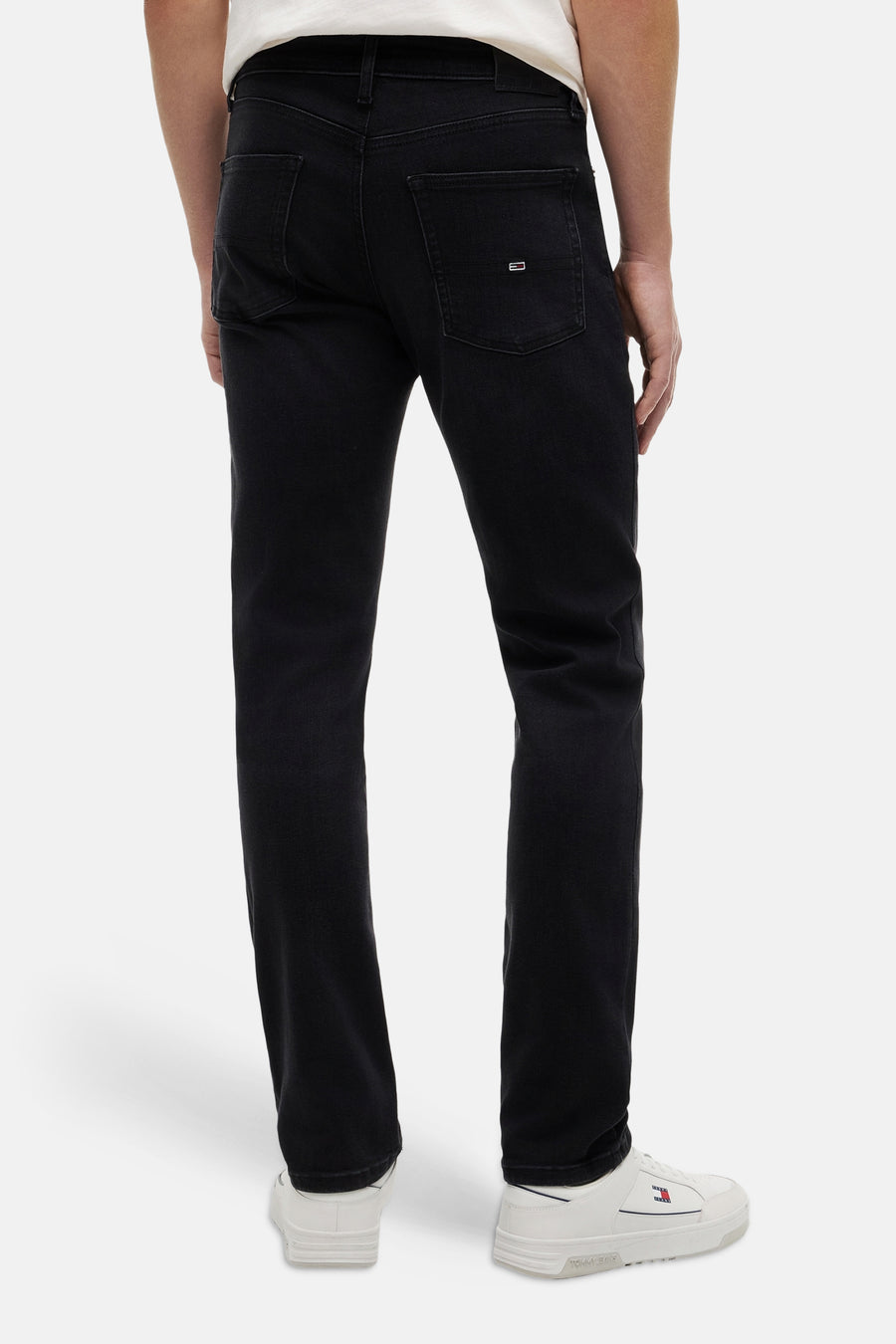 Jeans slim noir de TOMMY JEANS, vue arrière, avec petit logo drapeau sur la poche arrière droite, porté avec baskets et t-shirt blanc.