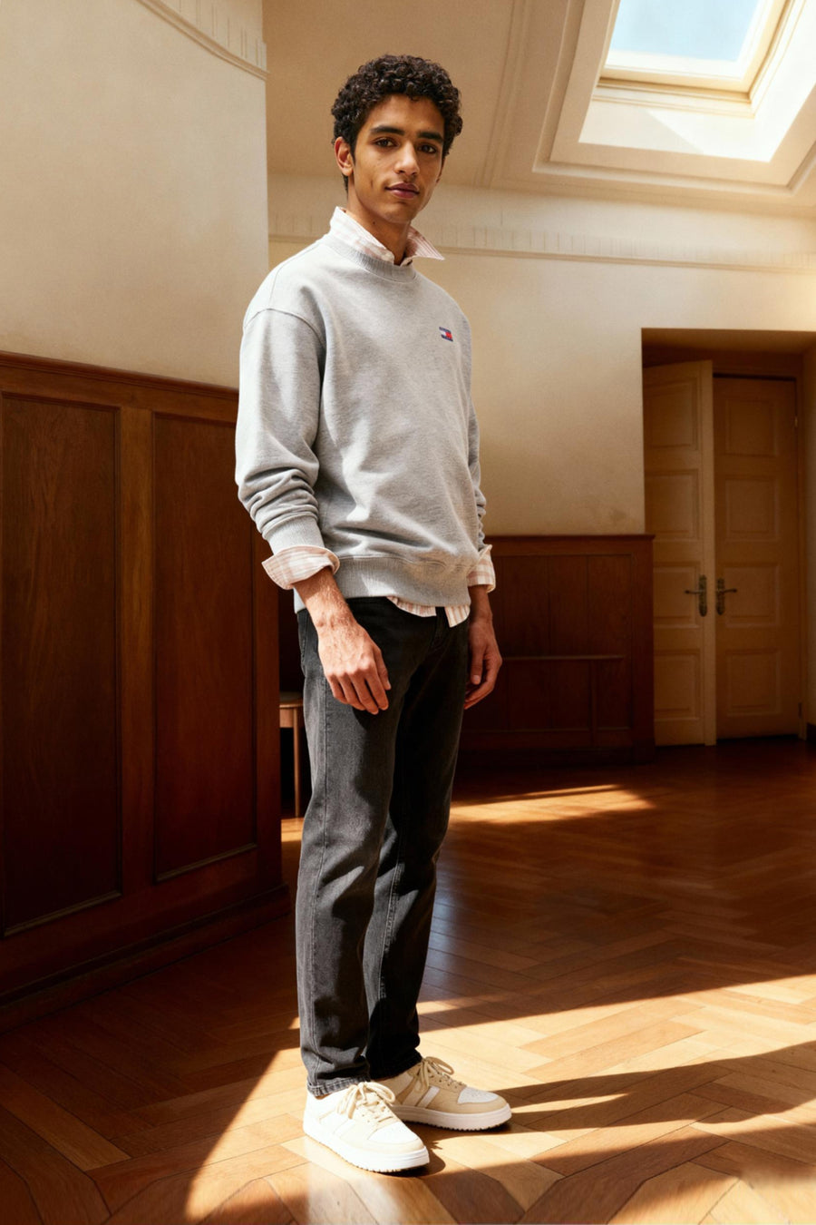 Jean slim noir en denim de Tommy Jeans, porté avec un pull gris et des baskets claires dans une pièce ensoleillée.
