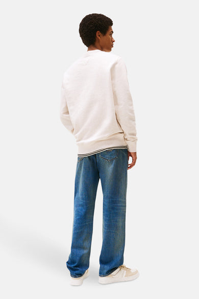 Sweat beige de TOMMY JEANS, avec un ourlet rayé, porté avec un jean bleu et des baskets crème.
