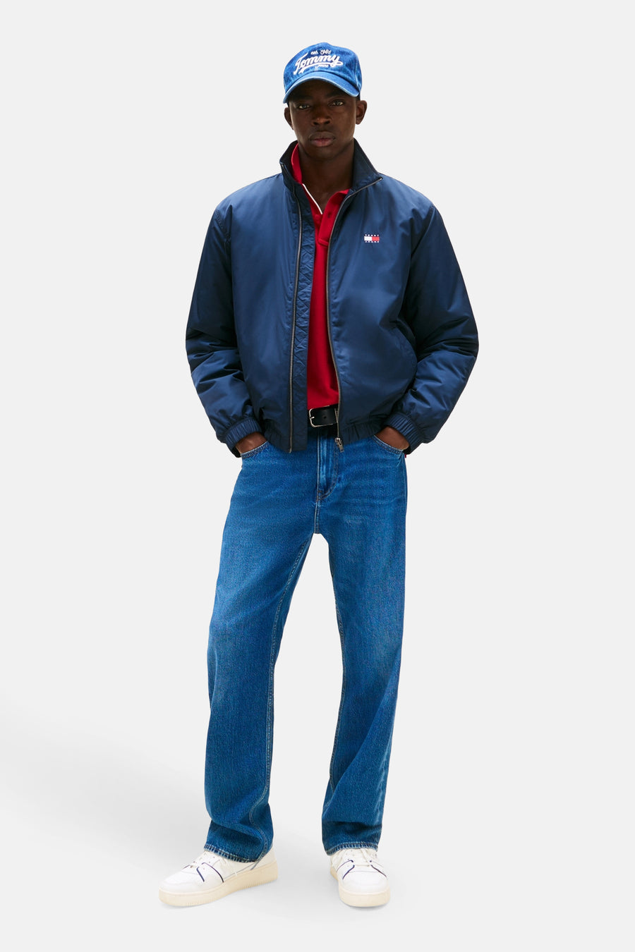 Imperméable bleu de TOMMY JEANS avec fermeture éclair portée avec polo rouge, jeans et casquette.