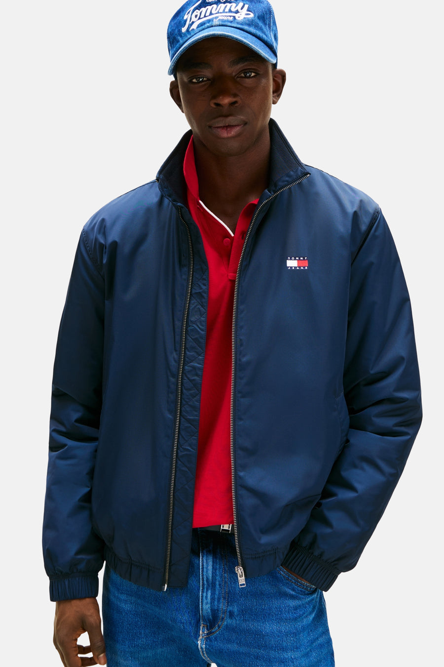Imperméable bleu de Tommy Jeans, porté avec un polo rouge et une casquette en denim.