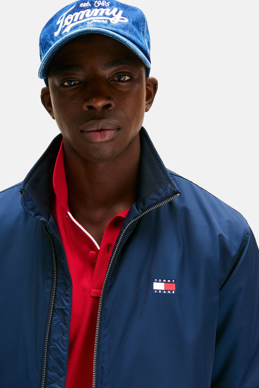 Imperméable bleu marine de TOMMY JEANS, porté sur un polo rouge, avec une casquette denim.