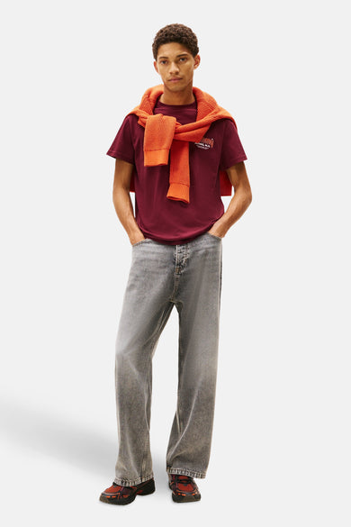 Rood T-shirt met korte mouwen van TOMMY JEANS, gedragen met een oranje trui geknoopt over de schouders en een grijze jeans.