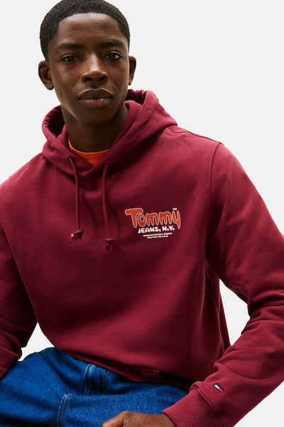 Sweat à capuche rouge de Tommy Jeans, avec cordon de serrage, porté avec un t-shirt orange et un jean bleu.