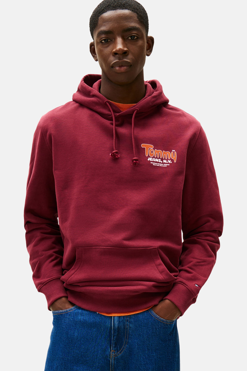 Bordeauxrode hoodie van Tommy Jeans, met trekkoord en logo op de voorkant, gedragen over een oranje T-shirt en een spijkerbroek.