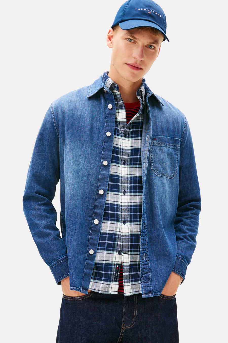 Chemise en jean bleu moyen de TOMMY JEANS, portée ouverte sur une chemise à carreaux et un t-shirt rayé, casquette bleue.