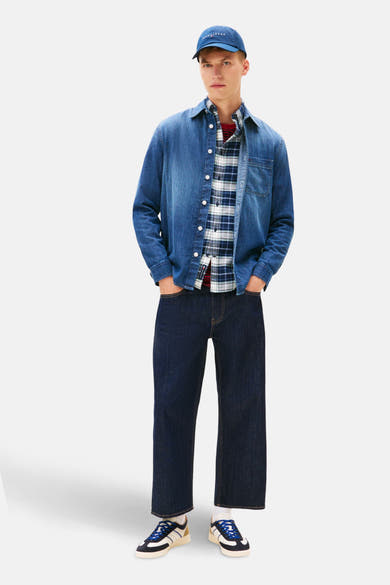 Chemise en jean bleu moyen de Tommy Jeans, portée ouverte sur une chemise à carreaux, avec un pantalon et une casquette bleu marine.