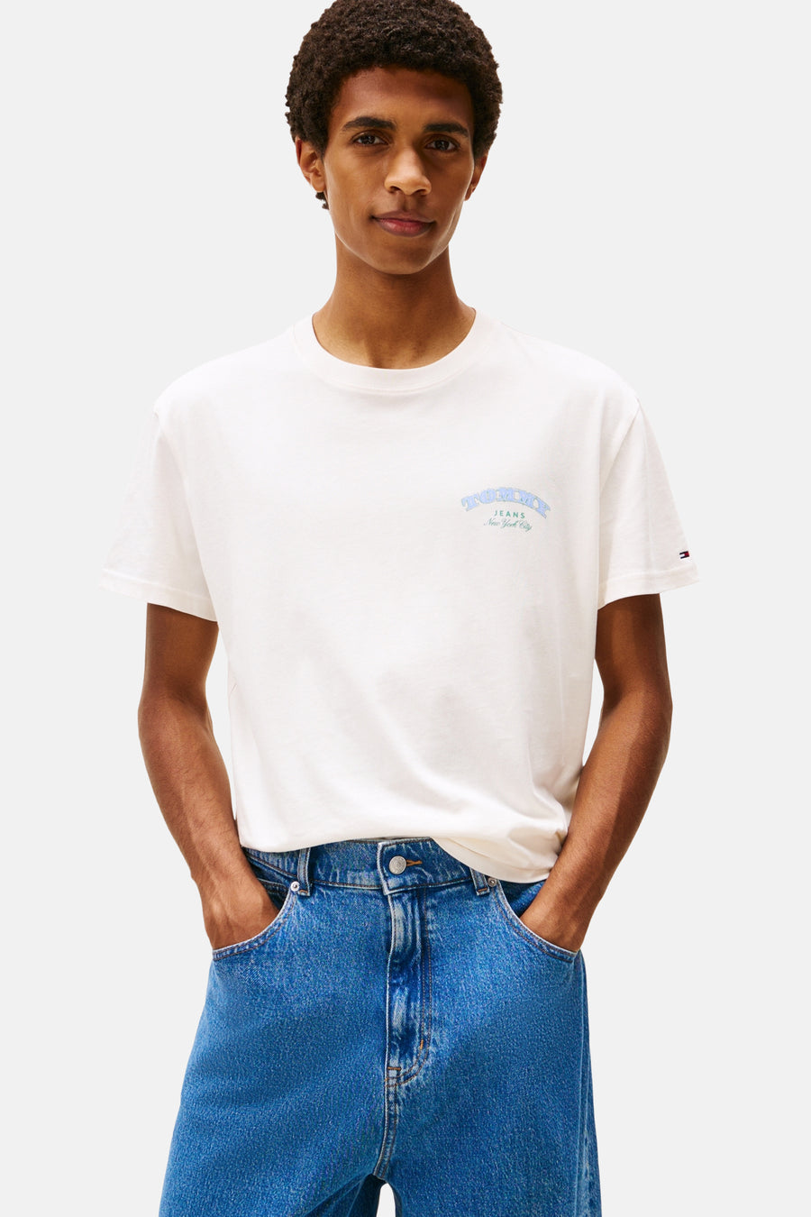 Ecru T-shirt met korte mouwen van TOMMY JEANS, met meerkleurige tekst op de rug, gedragen met een wijde blauwe jeans.