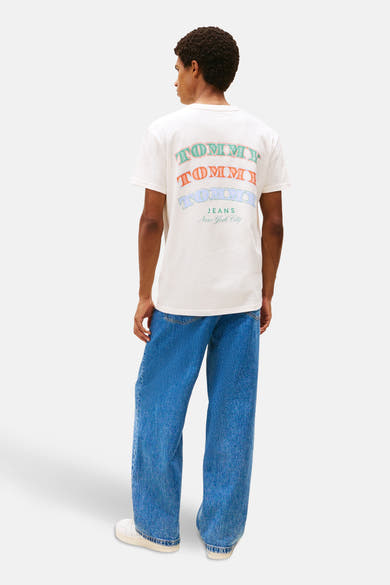 T-shirt à manches courtes écru de TOMMY JEANS, avec texte multicolore au dos, porté avec un jean bleu ample.