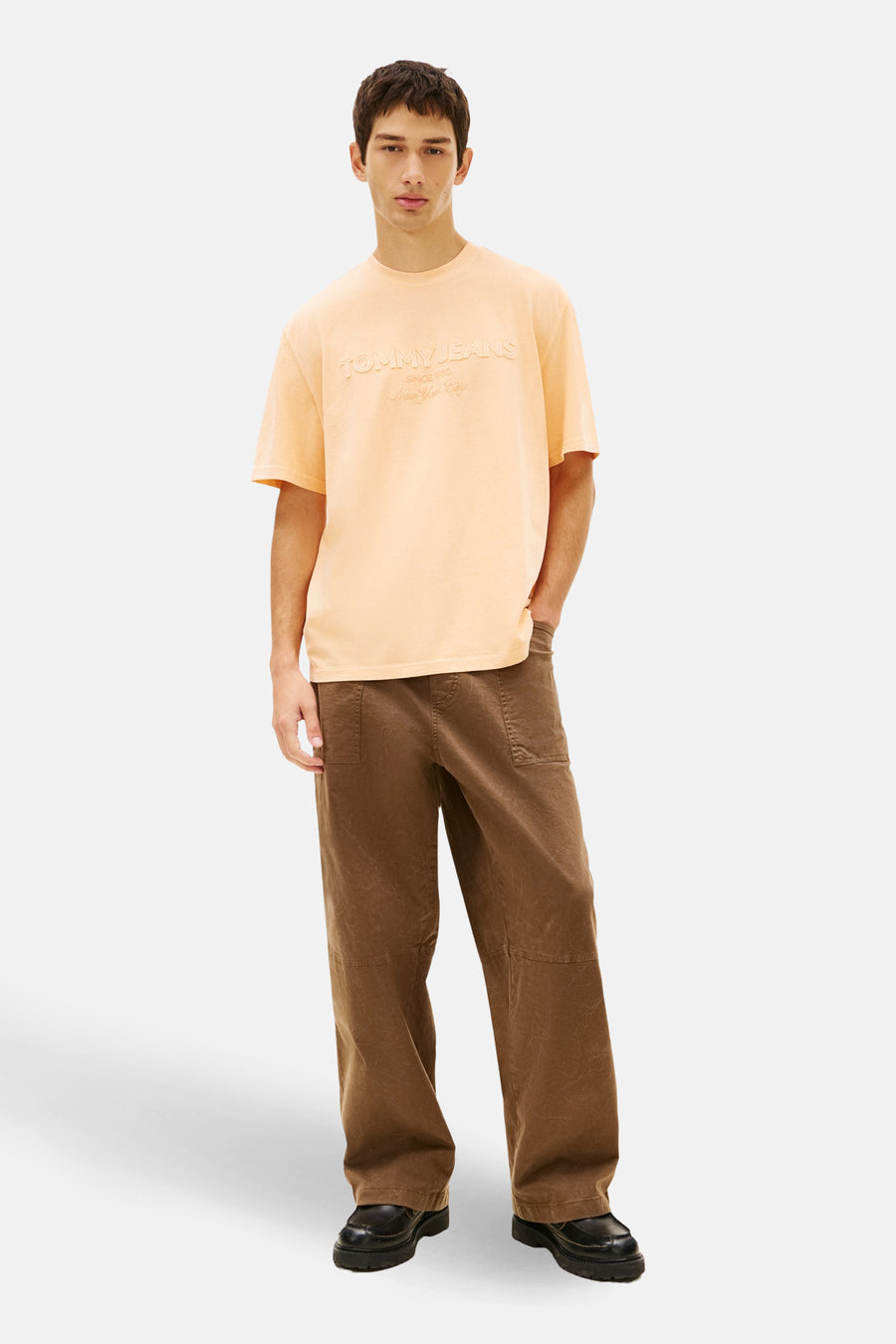 T-shirt orange à manches courtes de TOMMY JEANS, avec inscription en relief sur le devant, porté avec un pantalon marron.