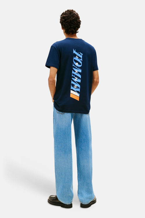 T-shirt bleu de TOMMY JEANS, avec un imprimé vertical sur le dos, porté avec un jean large et des chaussures noires.