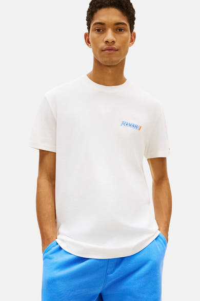 T-shirt blanc à manches courtes de TOMMY JEANS, avec logo imprimé sur la poitrine, porté avec un pantalon de survêtement bleu.