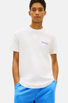 T-shirt blanc à manches courtes de TOMMY JEANS, avec logo imprimé sur la poitrine, porté avec un pantalon de survêtement bleu.