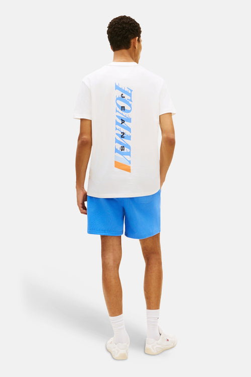 T-shirt blanc à manches courtes de TOMMY JEANS, avec imprimé vertical dans le dos, porté avec un short bleu.