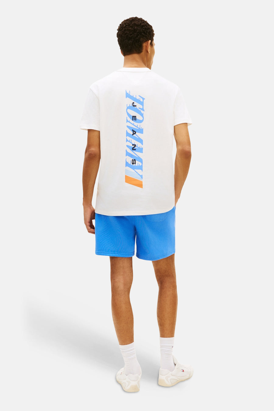 T-shirt blanc à manches courtes de TOMMY JEANS, avec imprimé vertical dans le dos, porté avec un short bleu.