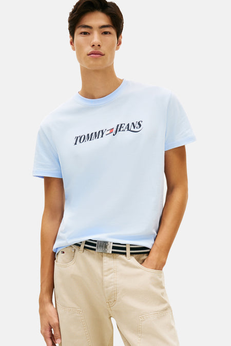 Lichtblauw T-shirt met korte mouwen van TOMMY JEANS, met logo op de voorkant, gedragen met een beige broek en bruine schoenen.