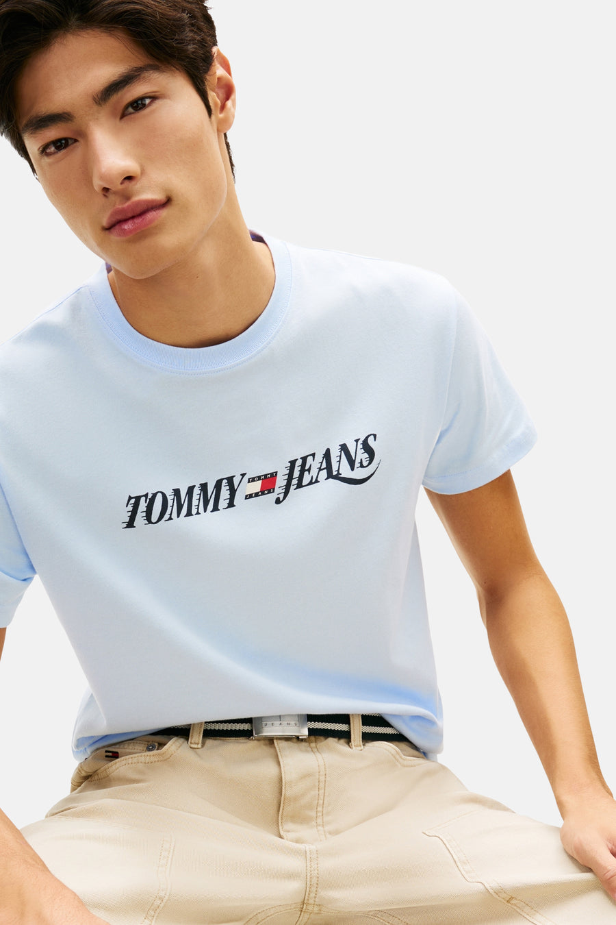 Lichtblauwe T-shirt van TOMMY JEANS, met logo op de voorkant, gedragen met een beige cargobroek en een gestreepte riem.