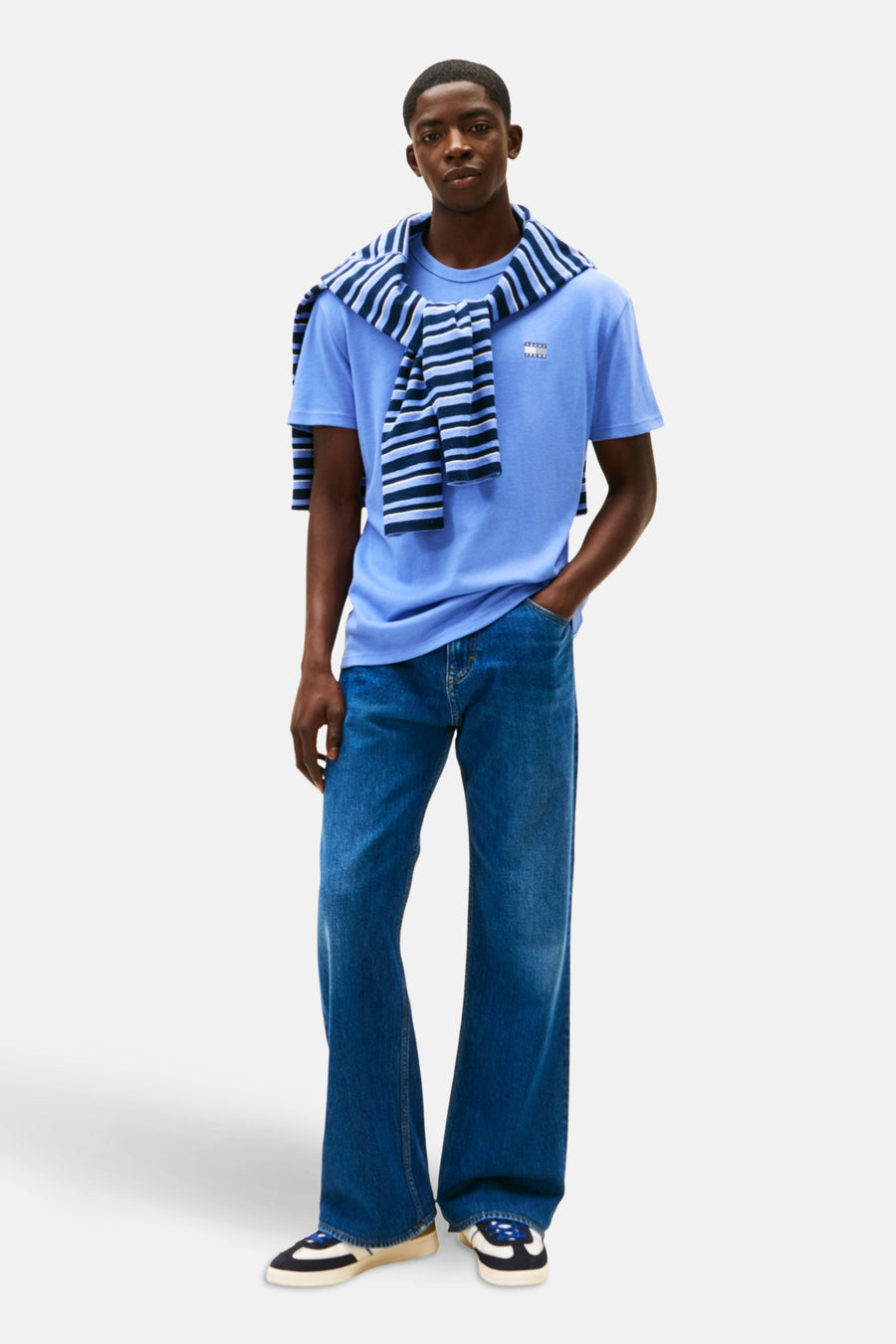 T-shirt à manches courtes bleu de TOMMY JEANS, avec un pull rayé noué autour du cou, porté avec un jean bleu ample.