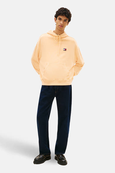 Sweat-shirt orange, par Tommy Jeans, avec capuche, poche kangourou et logo sur la poitrine, porté avec un jean bleu foncé et des chaussures noires.