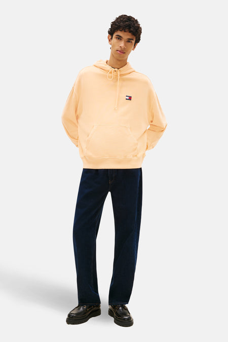 Sweat-shirt orange, par Tommy Jeans, avec capuche, poche kangourou et logo sur la poitrine, porté avec un jean bleu foncé et des chaussures noires.