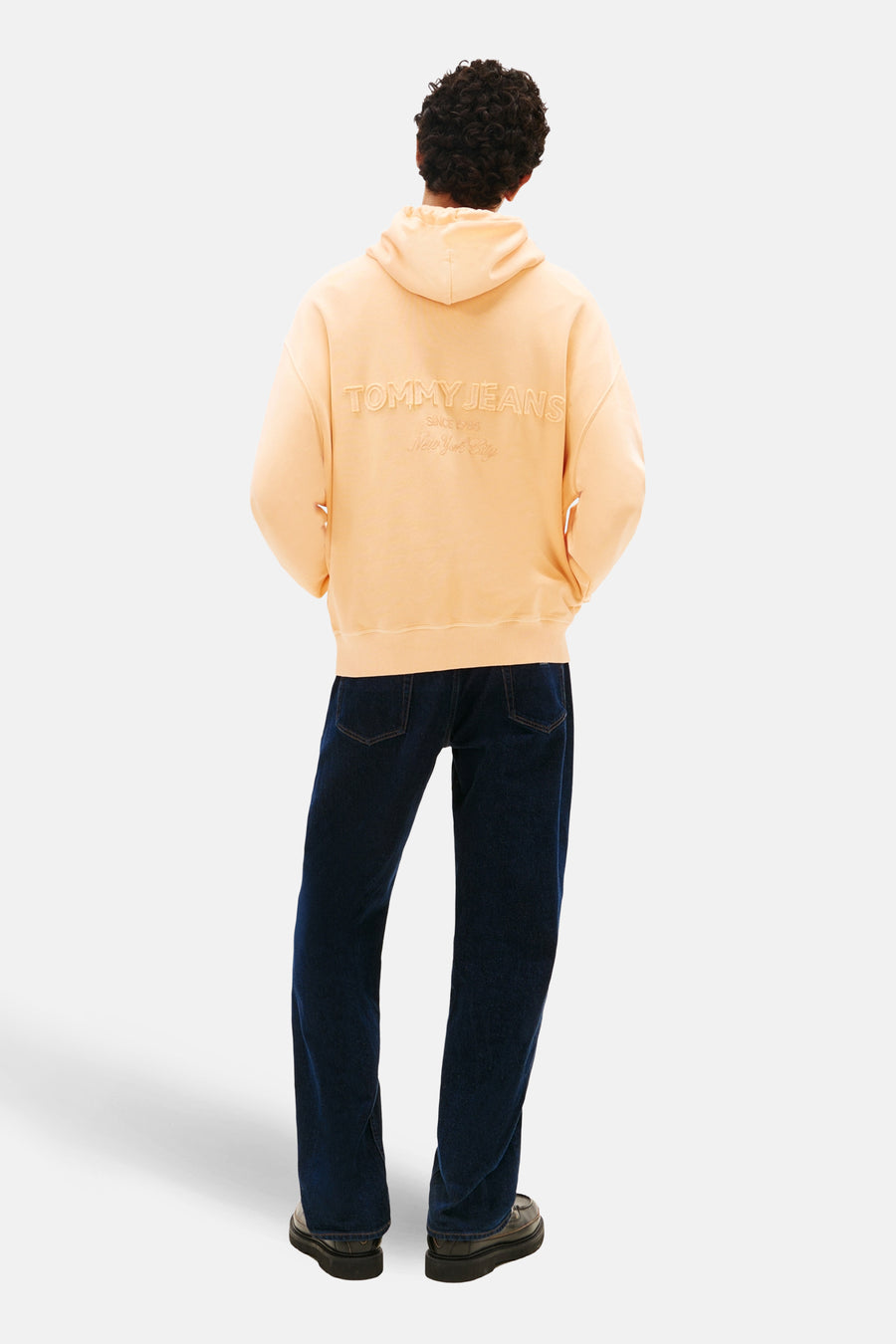 Oranje hoodie van TOMMY JEANS, met merkopdruk op de rug, gedragen met een donkerblauwe jeans en zwarte schoenen.