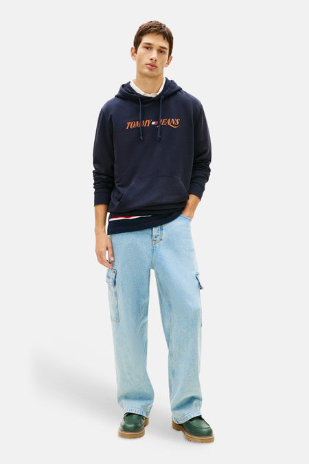 Sweat à capuche bleu de Tommy Jeans, porté avec un t-shirt rayé et un jean cargo délavé, complété par des chaussures vert foncé.