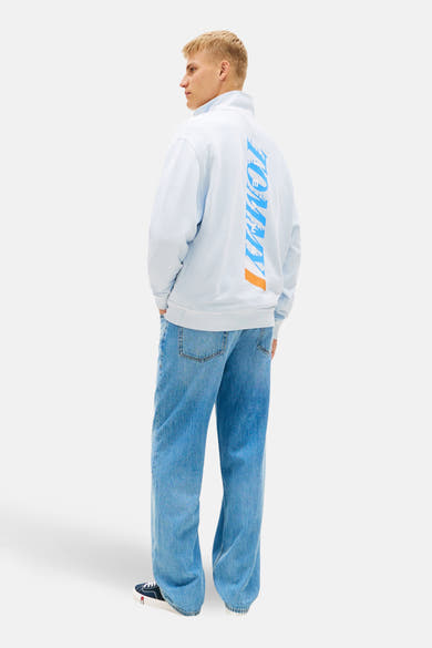 Blauwe sweater met opstaande kraag van TOMMY JEANS, met verticale opdruk op de rug, gedragen met een wijde jeans en sneakers.