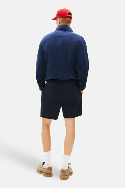 Short bleu de TOMMY JEANS, avec poches arrière et style décontracté, porté avec un pull bleu marine et des chaussures en daim.