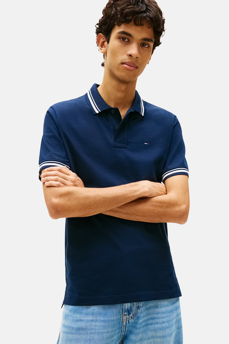Polo à manches courtes bleu marine de Tommy Jeans, avec des rayures blanches au col et aux manches, porté avec un jean clair.
