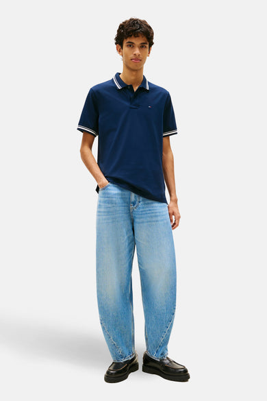 Polo à manches courtes bleu par Tommy Jeans, avec détails rayés blancs au col et aux manches, porté avec un jean ample et des mocassins noirs.
