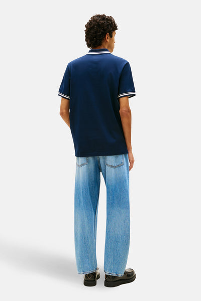 Polo à manches courtes bleu de TOMMY JEANS, avec rayures blanches au col et aux manches, porté avec un jean ample.
