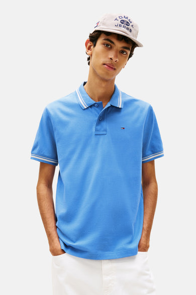 Polo bleu de TOMMY JEANS, avec col et manches rayés, porté avec une casquette.
