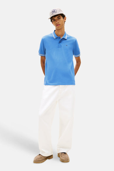 Polo bleu by TOMMY JEANS, avec col et manches à rayures blanches, porté avec un pantalon blanc et des chaussures marron.